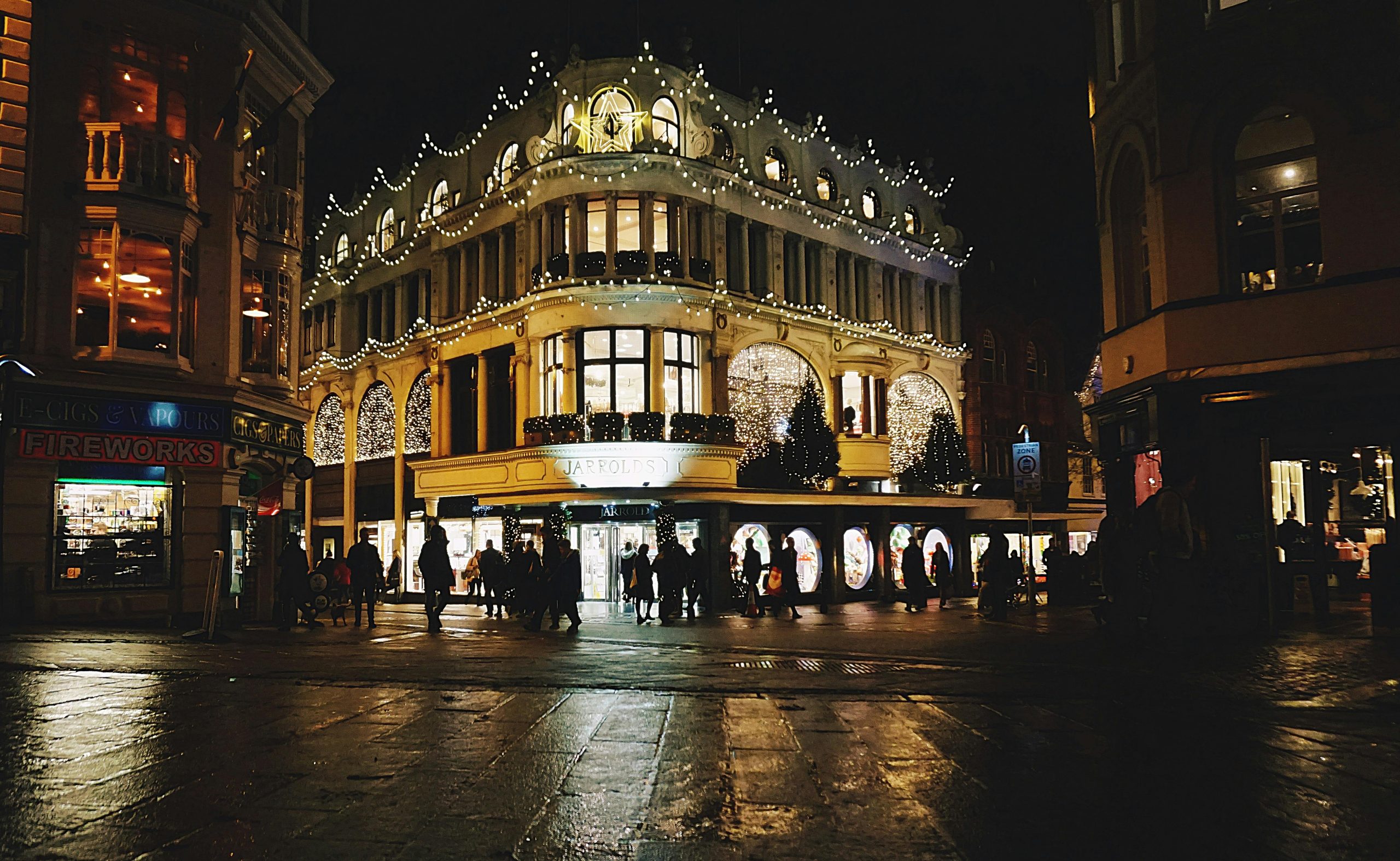 Jarrolds building illuminated with Christmas lights in Norwich at night, bustling with activity.